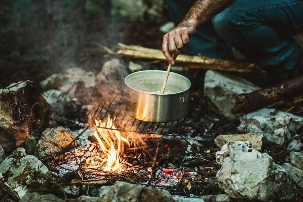 A person stirring a pot over a campfire, with the pot resting on a grill above the flames, surrounded by rocks.