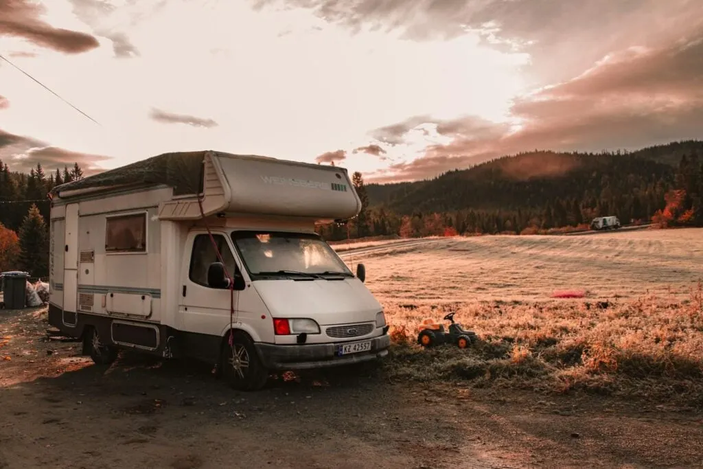 A camper van parked in a serene outdoor location with rolling hills and trees in the distance, captured during sunset. A small toy truck is placed next to the van, and another camper van is visible in the background.