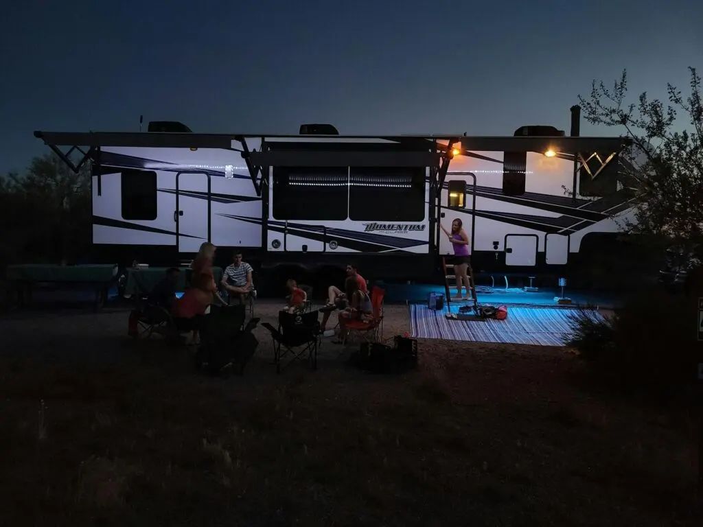 A nighttime scene of people gathered around an RV, illuminated by soft lighting. The group appears to be enjoying an outdoor gathering.