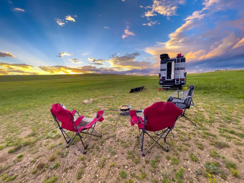 Foldable chairs arranged in a semi-circle around a small campfire, with a camper van in the background, set against a beautiful sunset in an open field.