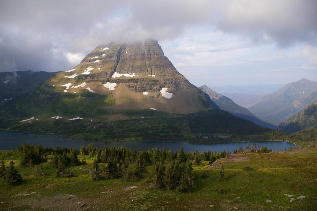 A majestic mountain with patches of snow on its steep slopes, towering over a serene lake and a dense forest of evergreen trees in Glacier National Park. The mountain's peak is partially shrouded in clouds.