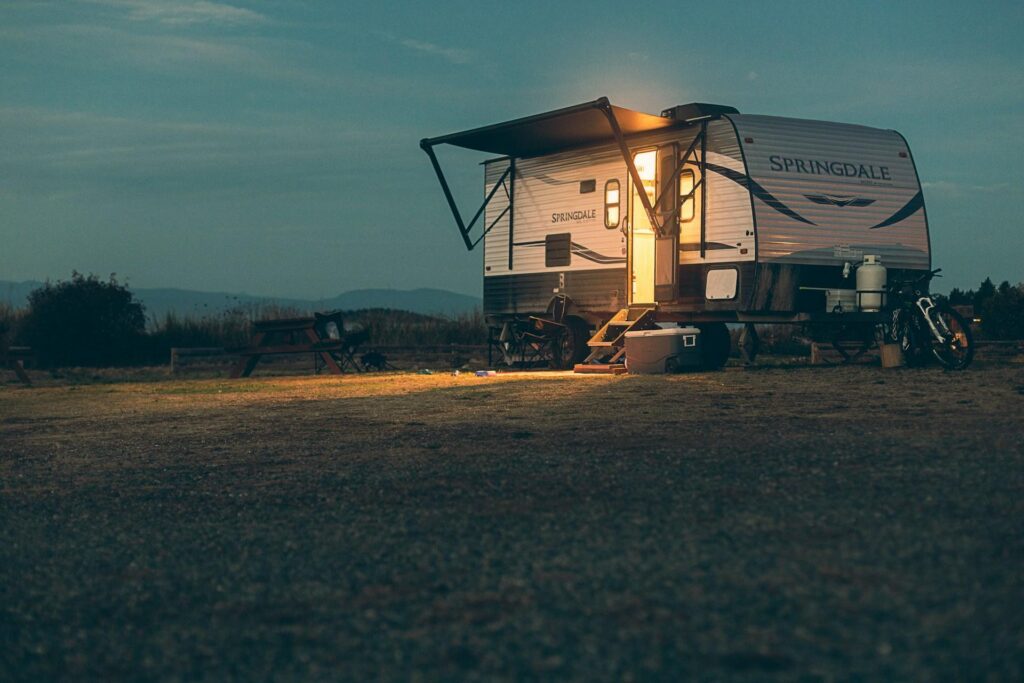 A well-lit camping trailer with an awning extended, parked at a campsite during twilight.