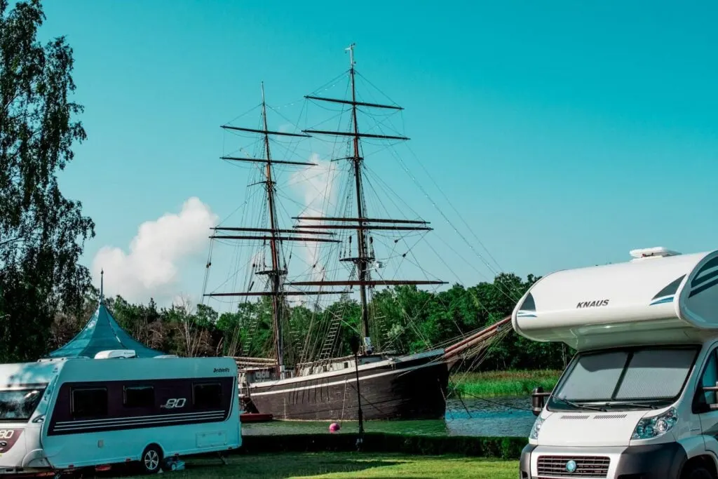 A campground with RVs parked near a body of water. A tall sailing ship with masts and rigging is visible in the background.