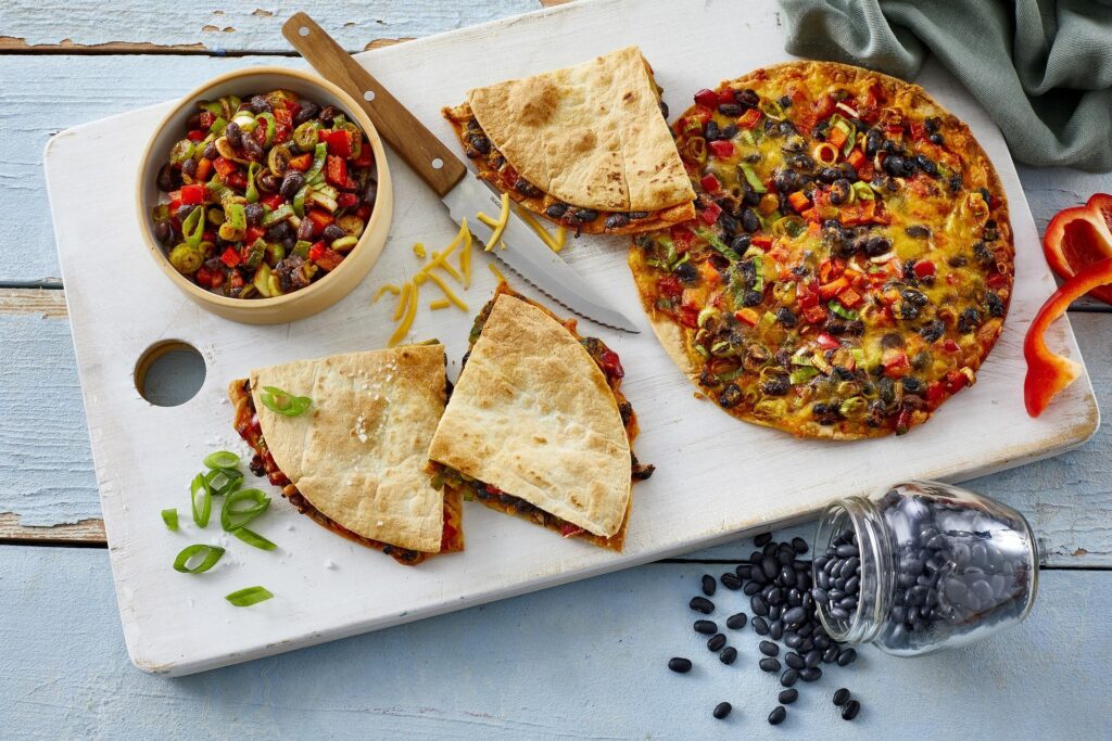 A top-down view of a white wooden board with a vegetable pizza, cut into slices, and small bowl of vegetable salad, surrounded by scattered black beans, chopped vegetables, and a knife.
