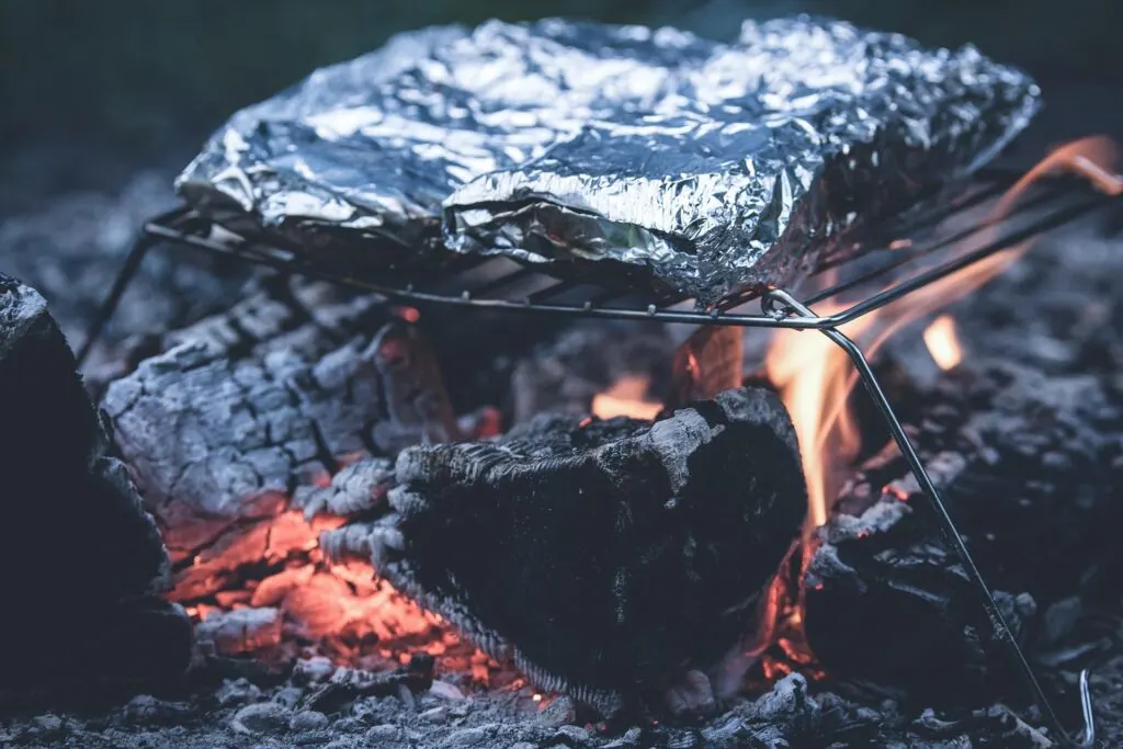 A foil-wrapped package sitting on a grill rack over glowing embers and open flames.