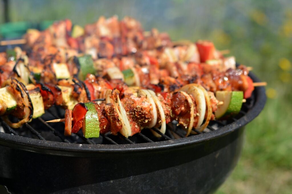 Close-up of sizzling skewers on a barbecue grill, loaded with marinated meat, zucchini slices, red bell peppers, and onions, all slightly charred from grilling. The background features an out-of-focus natural outdoor setting.