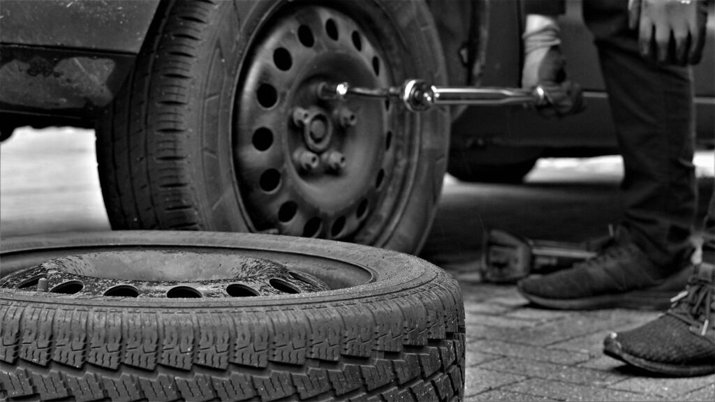 A black-and-white photo of a person changing a tire on a car, with a removed tire lying on the ground nearby.