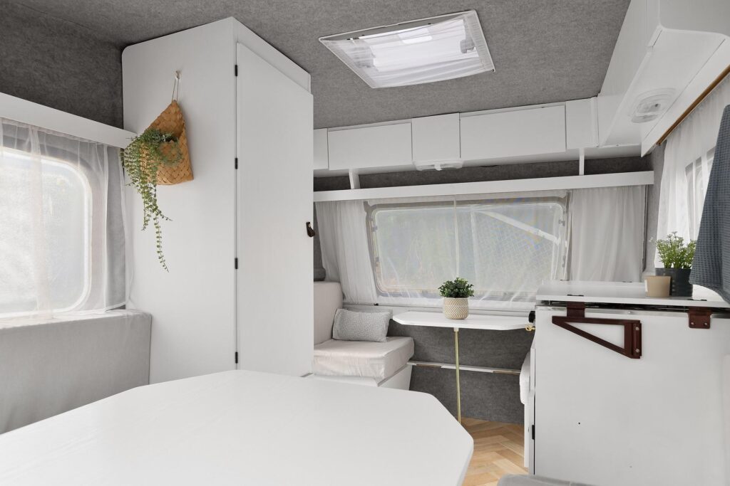 The interior of a camper van with minimalist white decor, featuring a small potted plant and a window with sheer curtains.