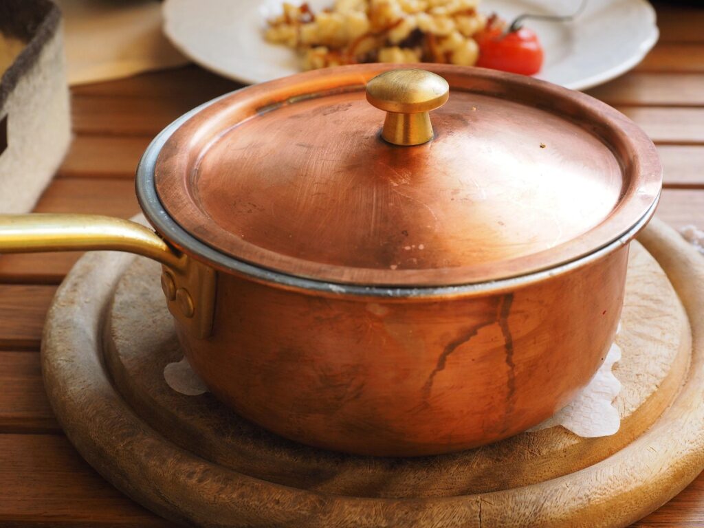 A copper pot with a lid on a wooden board, with a plate of food in the background.