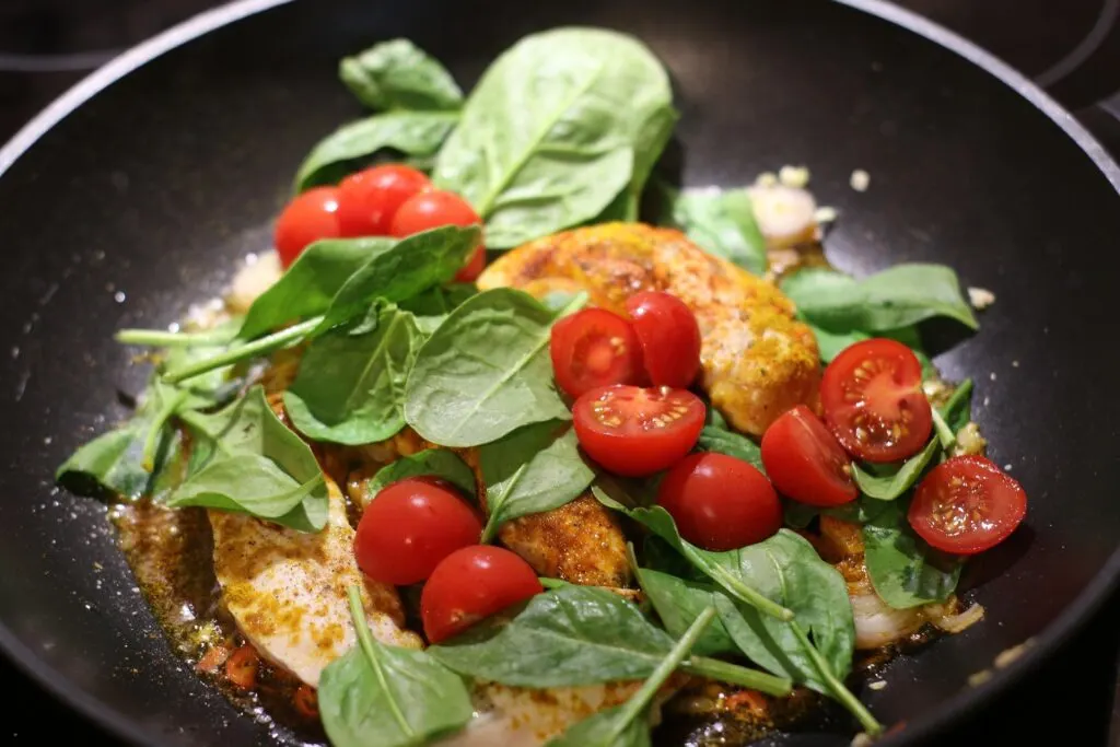 A close-up of a pan filled with chicken, cherry tomatoes, and fresh spinach leaves, being cooked.