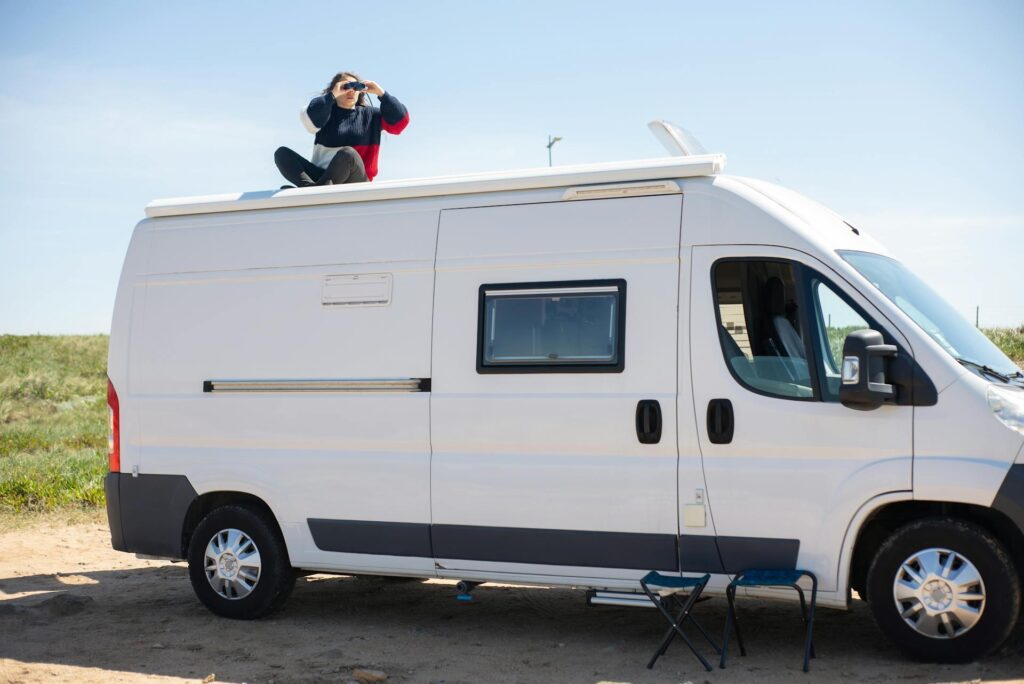 A person sitting on the roof of a white camper van, using binoculars to observe the surroundings at a scenic outdoor location. Two foldable chairs are placed beside the camper van.