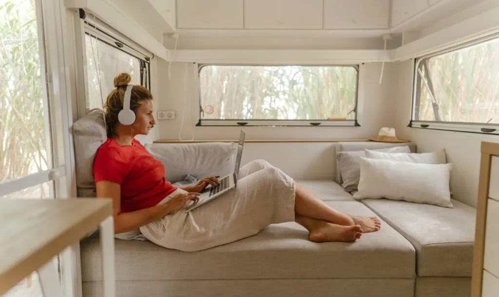 A woman in a red shirt and beige skirt is relaxing on a grey sofa inside a camper van, using a laptop while wearing white headphones. The space is bright and airy with windows on both sides, offering views of greenery outside.