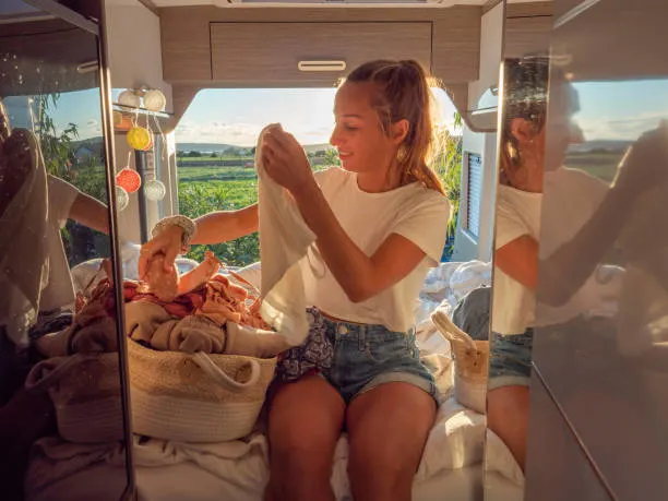 A young woman sitting on a bed inside a camper van, holding a piece of clothing and sorting through a basket of laundry. She is wearing a white T-shirt and denim shorts, illuminated by the warm sunlight streaming through the windows. The interior is cozy, with a view of the scenic landscape outside the windows.