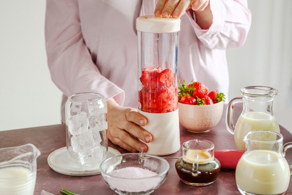 A woman in a pink shirt is using a blender to blend strawberries. The countertop in front of her  is filled with various ingredients, including a glass of ice cubes, a bowl of fresh strawberries, a small pitcher of milk, a smaller pitcher of cream, a glass cup filled with coffee, and a bowl of powdered mix.