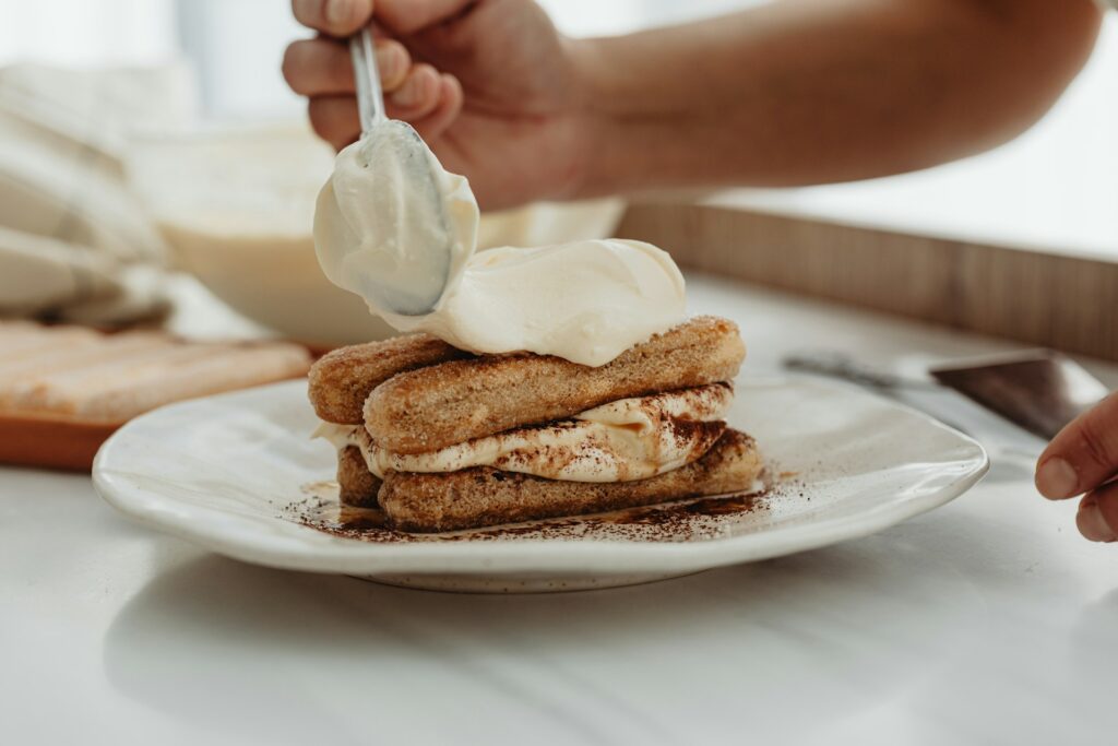 A person spooning cream onto a plate of layered tiramisu, with the dessert being carefully assembled on a white plate.