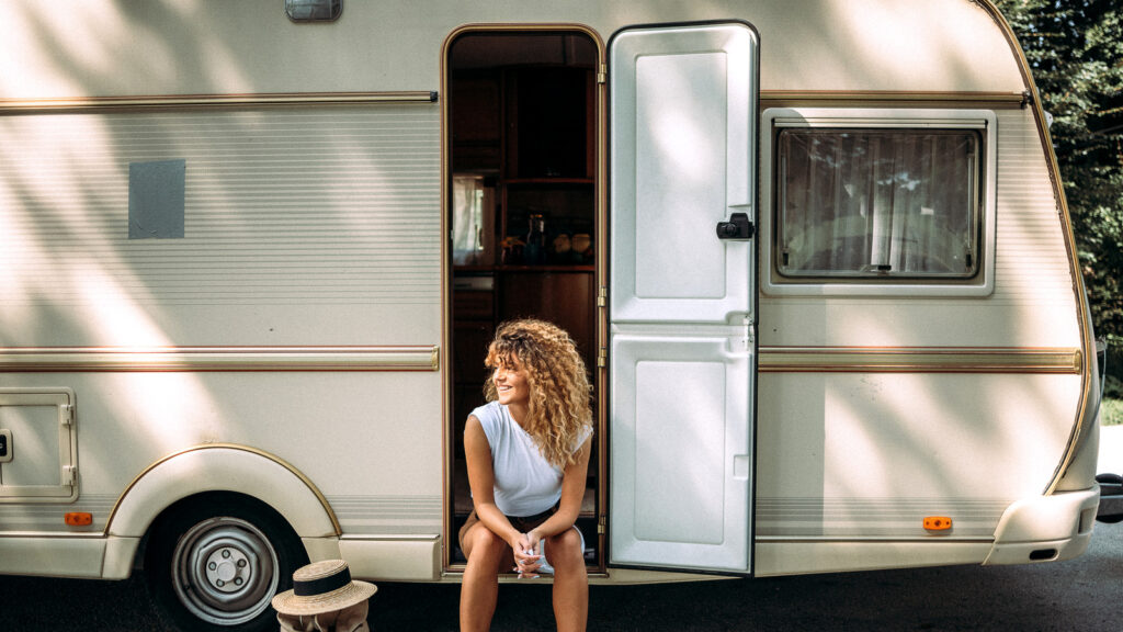 A person with curly hair sits on the steps of a parked camper van, enjoying the outdoors with a hat placed beside them.