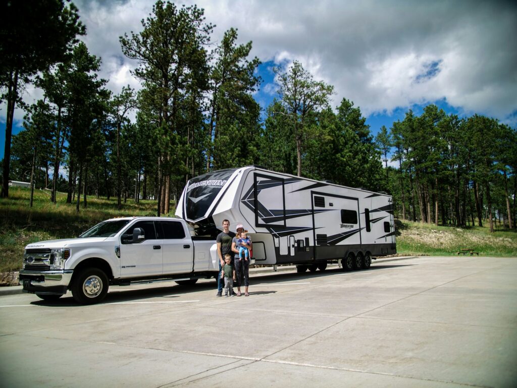 A family posing beside their large white truck and fifth-wheel RV trailer in a scenic, tree-lined parking area. The clear sky and dispersed clouds complement the lush, green trees in the background, creating a perfect setting for a road trip or camping adventure.