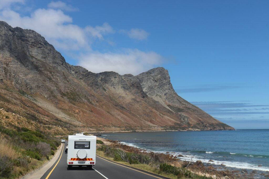 A white RV traveling along a scenic coastal road with towering cliffs on one side and the ocean on the other, under a clear blue sky.