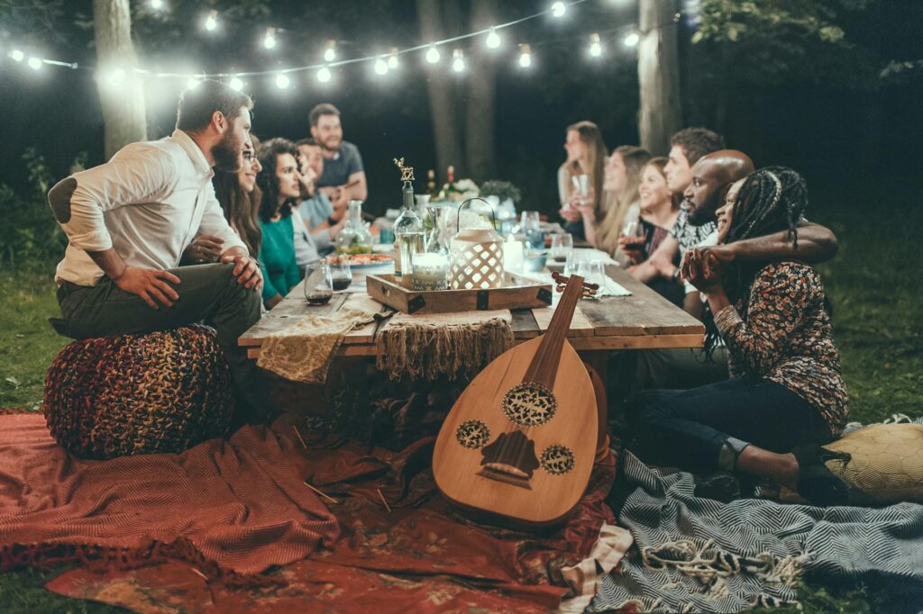 A group of friends at an outdoor dinner party, sitting around a table decorated with string lights and lanterns, enjoying a meal and drinks. A guitar rests against the table