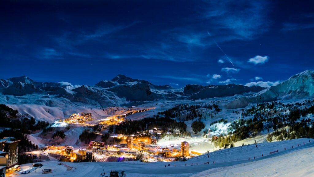 A stunning night view of a snowy mountain resort illuminated with warm lights. The resort buildings glow against the dark blue sky, which is dotted with stars and wispy clouds. The surrounding landscape is blanketed in snow, with majestic mountain peaks in the background. The ski slopes and runs are visible, creating a picturesque alpine scene.