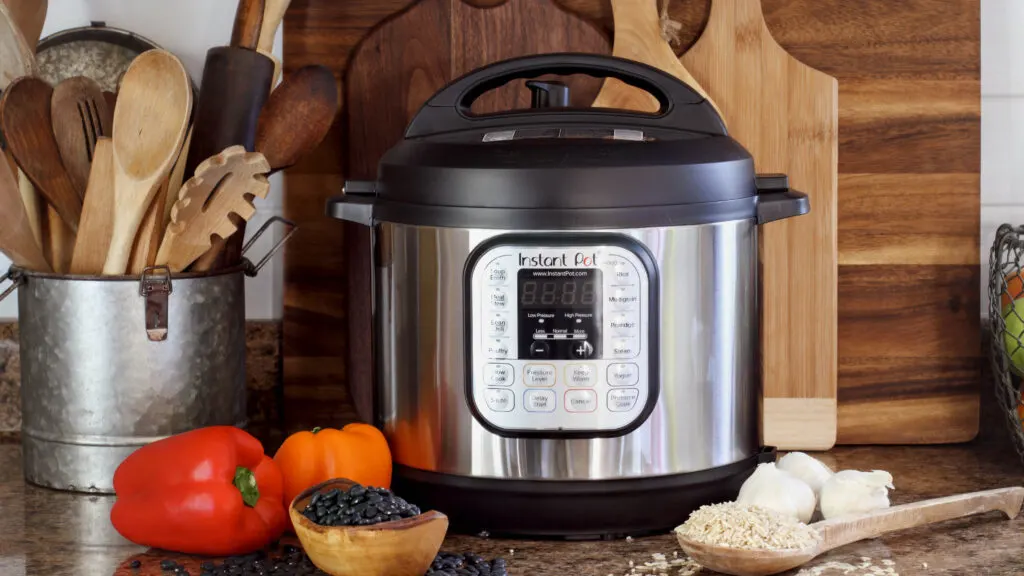 A kitchen countertop featuring an Instant Pot pressure cooker surrounded by cooking ingredients and utensils. To the left, a metal canister holds various wooden utensils. In front of the Instant Pot, there are a red and an orange bell pepper, a small bowl of black beans, a wooden spoon filled with rice and garlic bulbs. Wooden cutting boards are neatly arranged in the background.