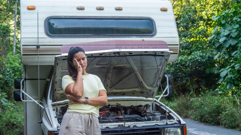 A woman standing in front of a motorhome with the hood open, appearing concerned or frustrated, surrounded by greenery.