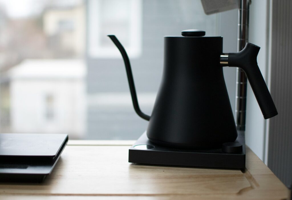 A black electric gooseneck kettle sitting on a wooden surface near a window, with natural light illuminating it. The background shows a blurred view of neighboring buildings and a closed laptop on the left side of the table.