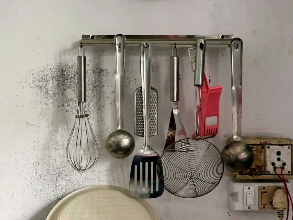 A set of stainless steel kitchen utensils hanging on a wall-mounted rack, with a whisk, ladle, spatula, strainer, and grater.