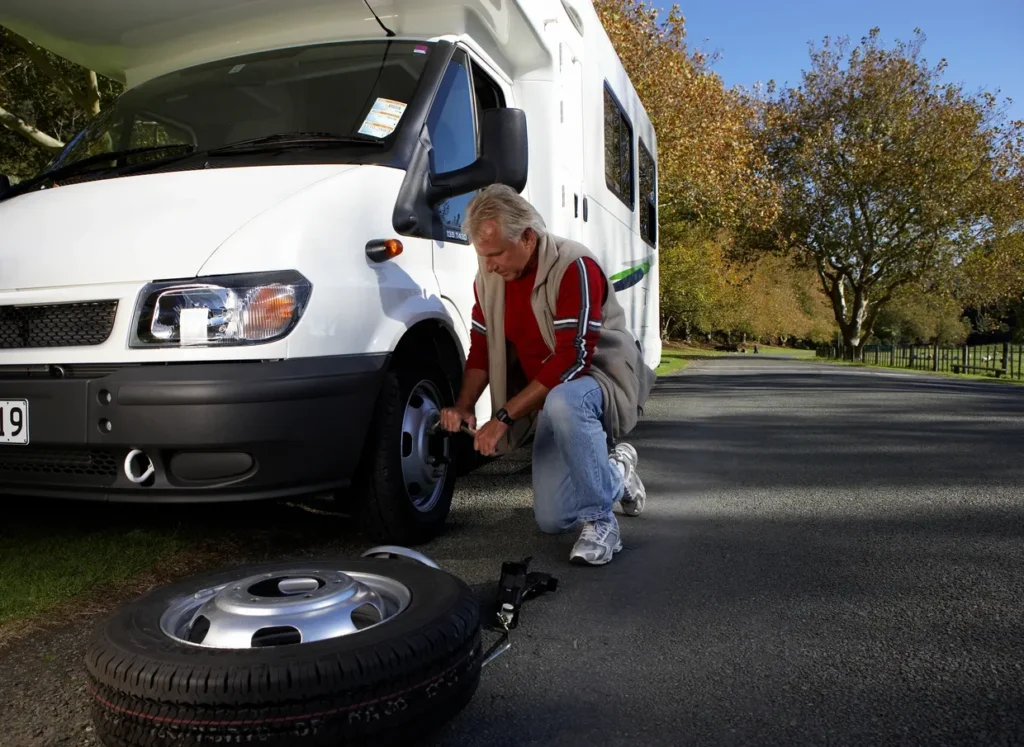 A man is repairing a tire on his RV. The RV is standing picturesque road surrounded by the autumn trees.