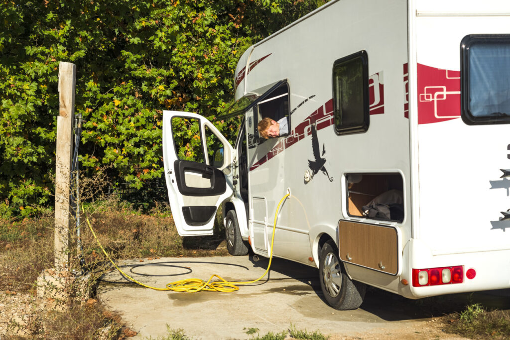 A camper van parked on a concrete pad next to a wooded area with its driver-side door open and a yellow hose attached to an external hookup. A man peeks out from the passenger window, likely checking the connection. The lush green trees in the background create a serene, natural setting.