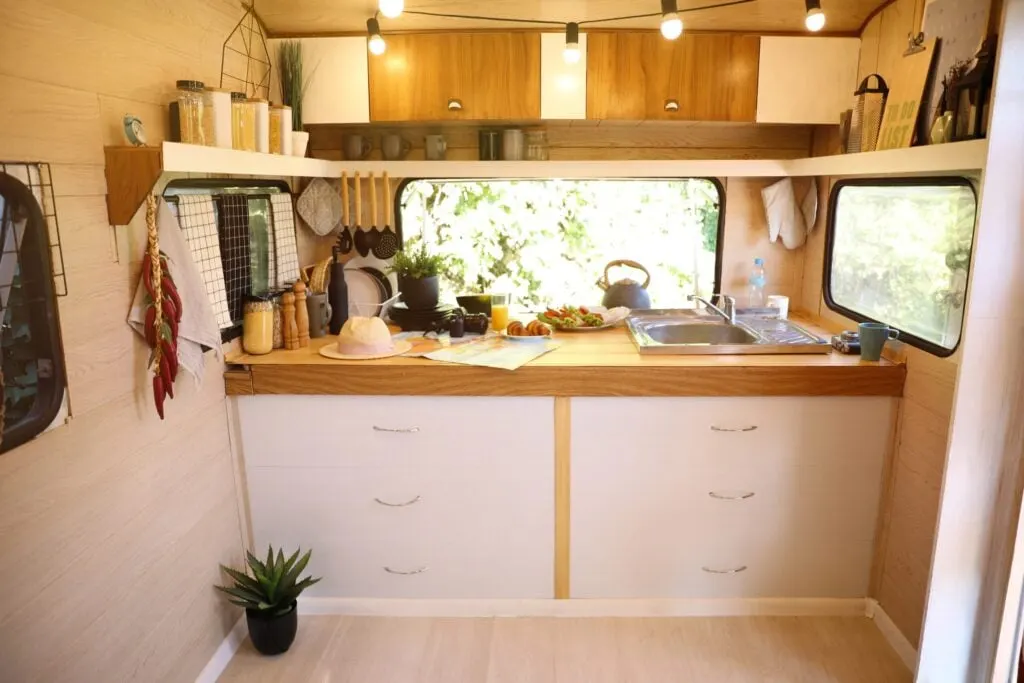 A cozy camper van kitchen with a wooden countertop and white cabinets. The countertop features various kitchen items, including a kettle, a cutting board with sliced vegetables, a glass of orange juice, a straw hat, and a map. Warm lighting hangs from the ceiling, enhancing the inviting atmosphere.