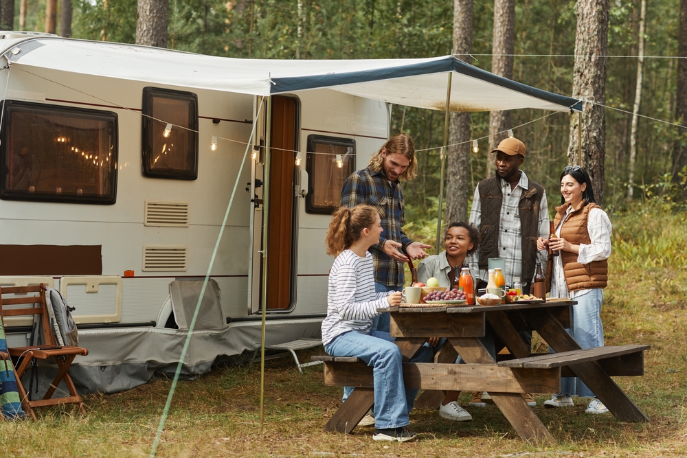 A group of friends enjoying an outdoor meal at a picnic table next to a camper van, set up in a wooded area with string lights hanging overhead.