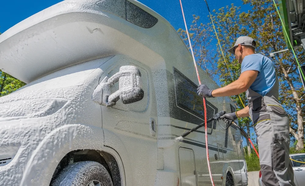 A person washing a motorhome with a high-pressure hose, covering it in soap foam, on a bright sunny day.