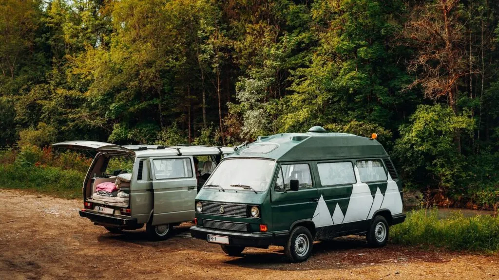 Two camper vans parked on a dirt area near a forest, with their back doors open, revealing camping gear and bedding inside.