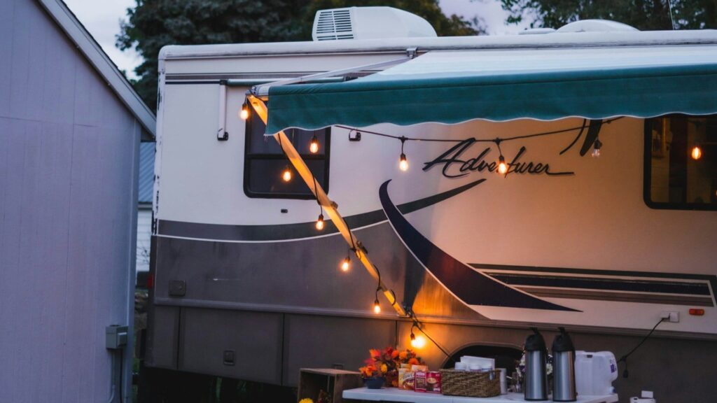 The side view of a camper van with a green awning extended, creating a shaded area. String lights are hung around the awning, illuminating the evening scene. Below the awning, there is a small outdoor setup with a table holding various items, including a fall-themed centerpiece and drink dispensers. The word "Adventurer" is written on the side of the camper van.