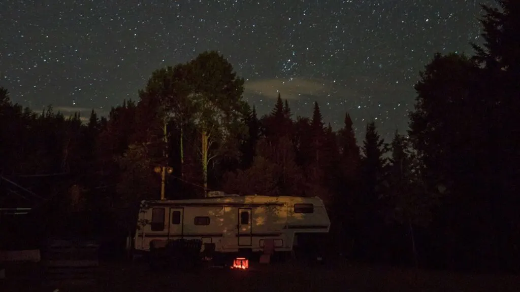 A camper van parked in a forest clearing under a star-filled night sky, with a small campfire glowing nearby.