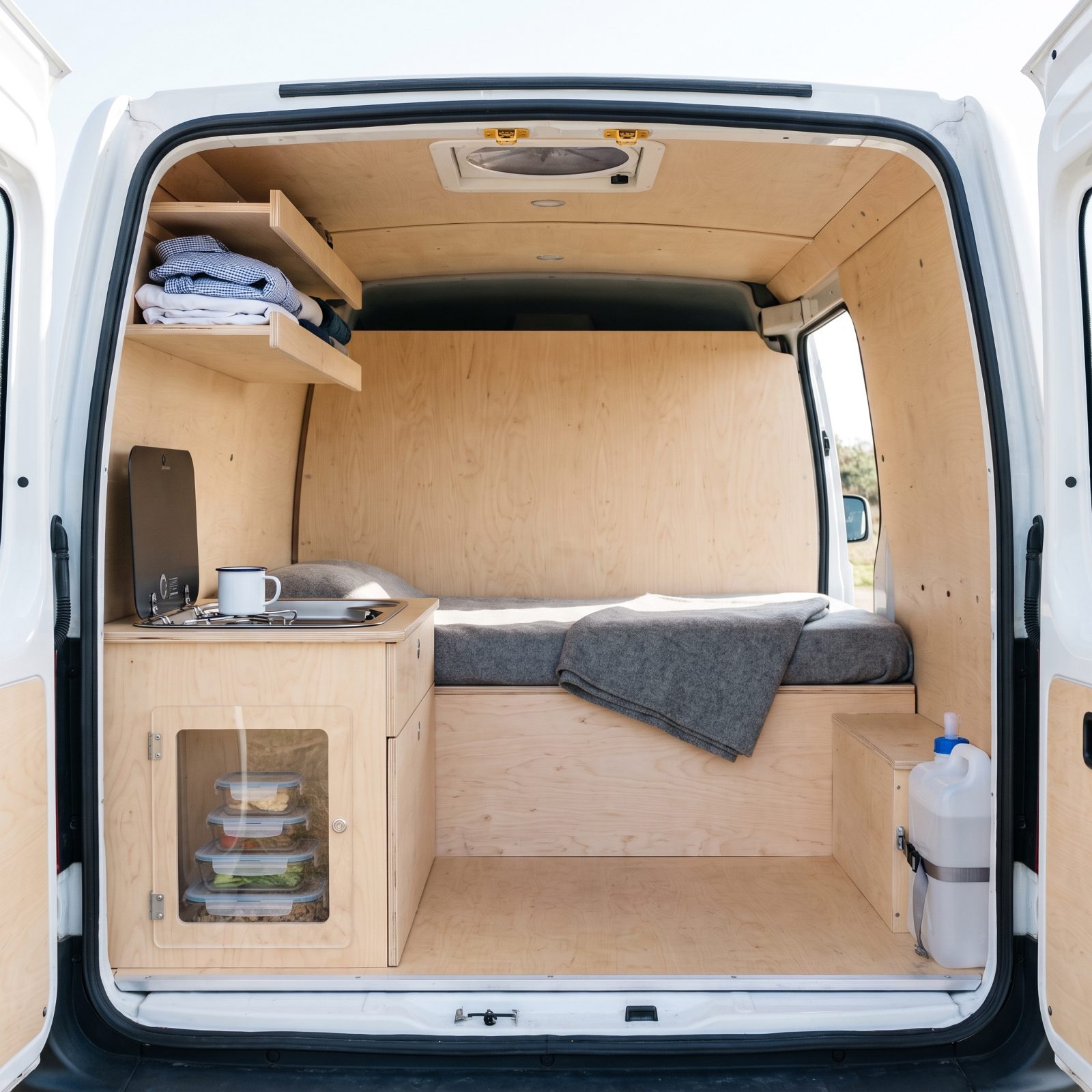 Interior of a camper van conversion with a single-burner galley, one shelf of folded shirts, and a bed platform across the